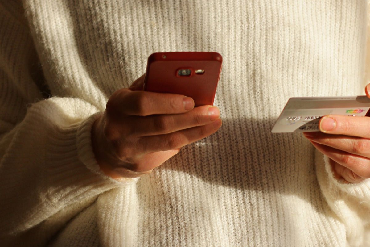 Manos sosteniendo un celular y una tarjeta, con un suéter color crema de fondo